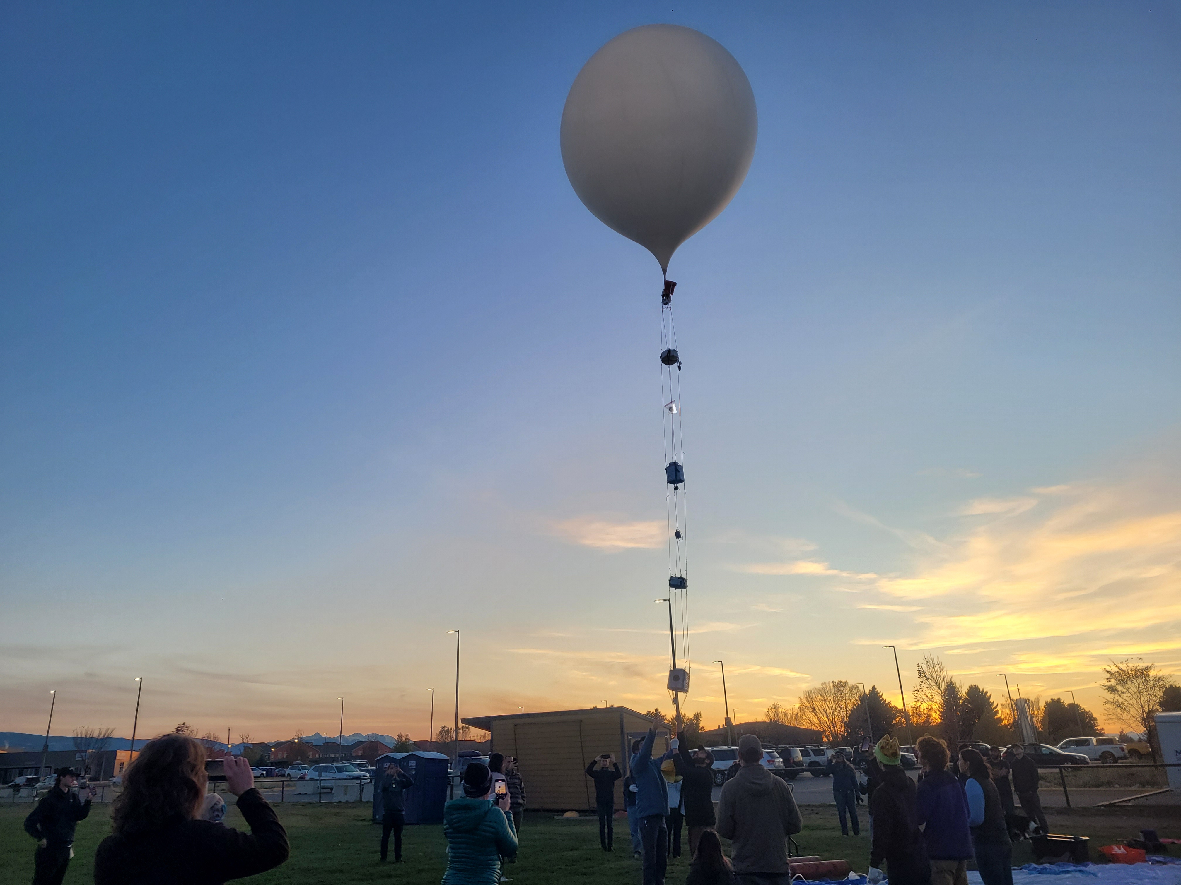 A balloon with many payloads surrounded by a crowd of people at dusk